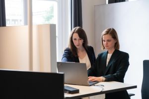 Zwei Frauen stehen nebeneinander an einem Schreibtisch vor einem Laptop
