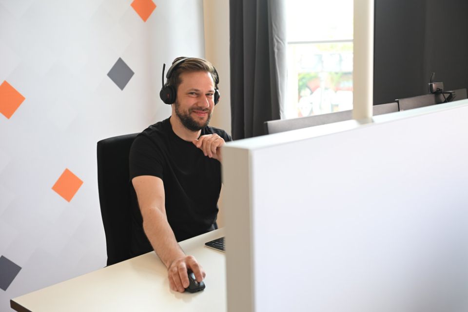 Eine Person sitzt am Schreibtisch vor einem PC und lächelt in die Kamera